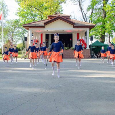Szerokie ujęcie grupy kilkunastu mażoretek w różnym wieku, ubranych w granatowo-pomarańczowe stroje i furażerki. Dziewczęta tańczą na dużym placu przed amfiteatrem w słoneczny dzień.