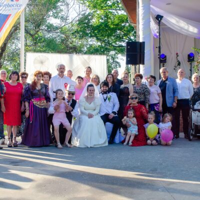 "Zbiorowe zdjęcie grupy około dwudziestu osób w różnym wieku, pozujących z parą młodą siedzącą w centrum. Panna młoda w białej sukni, pan młody w białym garniturze z czarnymi klapami. W tle biała dekoracja i scena z muzykami. Widać flagę z napisem 'Grębocice Przyjazna Gmina'. Z przodu siedzą małe dzieci z kolorowymi balonami. Jeden mężczyzna gra na akordeonie."