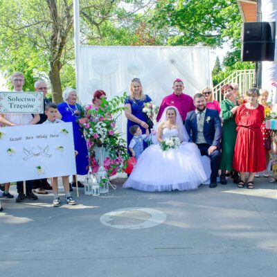 "Grupa osób pozuje wokół pary młodej siedzącej na krzesłach. Panna młoda w białej sukni bez ramiączek, pan młody w ciemnoniebieskim garniturze i jasnej kamizelce. Za parą stoi mężczyzna w fuksjowym stroju duchownym z piuską. Po lewej stronie młode osoby trzymają dwie tablice, na jednej z nich napis 'Sołectwo Trzęsów', a na drugiej 'Szczęść Boże Młodej Parze'."