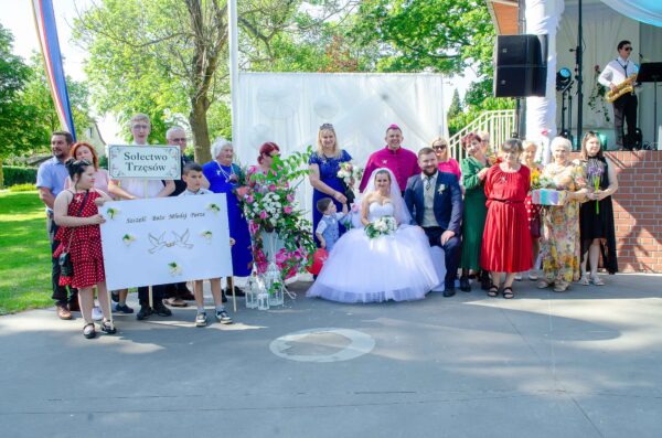 "Grupa osób pozuje wokół pary młodej siedzącej na krzesłach. Panna młoda w białej sukni bez ramiączek, pan młody w ciemnoniebieskim garniturze i jasnej kamizelce. Za parą stoi mężczyzna w fuksjowym stroju duchownym z piuską. Po lewej stronie młode osoby trzymają dwie tablice, na jednej z nich napis 'Sołectwo Trzęsów', a na drugiej 'Szczęść Boże Młodej Parze'."