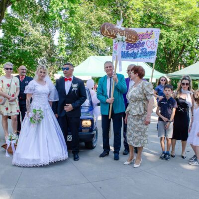 "Grupa osób i para młoda pozują na tle zielonych drzew i namiotów. Panna młoda w białej sukni, pan młody w garniturze z czerwoną muszką i okularach, stoją w centrum obok fragmentu ciemnoniebieskiego samochodu ozdobionego balonami. Osoby trzymające tablice 'Kwielice' oraz 'Szczęść Boże...' stoją po prawej stronie pary młodej. Widać też grupę dzieci i młodzieży."