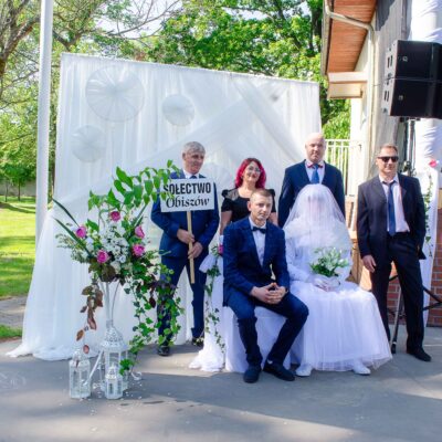 "Mniejsza, kilkuosobowa grupa pozująca wokół siedzącej pary. Panna młoda w całości zasłonięta długim, gęstym białym welonem i białym okryciem, trzyma bukiet kwiatów. Pan młody siedzi obok w ciemnoniebieskim garniturze w kratę. Jeden z mężczyzn stojących za parą trzyma białą tablicę z czarnym napisem: 'SOŁECTWO Obiszów'."