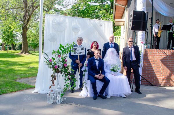 "Mniejsza, kilkuosobowa grupa pozująca wokół siedzącej pary. Panna młoda w całości zasłonięta długim, gęstym białym welonem i białym okryciem, trzyma bukiet kwiatów. Pan młody siedzi obok w ciemnoniebieskim garniturze w kratę. Jeden z mężczyzn stojących za parą trzyma białą tablicę z czarnym napisem: 'SOŁECTWO Obiszów'."