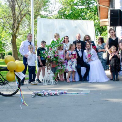 "Grupa osób pozuje wokół siedzącej pary młodej na tle dekoracji. Panna młoda w białej sukni z bukietem, pan młody w ciemnym garniturze. Za parą stoi mężczyzna w czarnej sutannie (duchowny). Na pierwszym planie po lewej stronie widać czarny rower ozdobiony żółtymi balonami, ciągnący rząd pustych puszek."