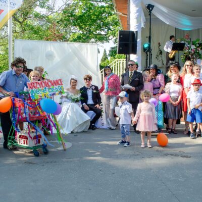 "Grupa osób w zróżnicowanych, kolorowych strojach (część w stylu retro/przerysowanym) pozuje wokół pary siedzącej w centrum. Panna młoda w sukni z odkrytymi ramionami, pan młody w ciemnym garniturze i okularach. Po lewej stronie mężczyzna w peruce trzyma koszyk zakupowy z dekoracją i tablicą 'KRZYDŁOWICE Wesele Lat 90'."