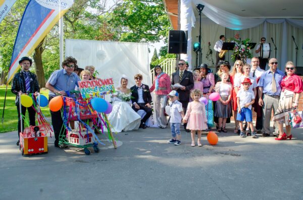 "Grupa osób w zróżnicowanych, kolorowych strojach (część w stylu retro/przerysowanym) pozuje wokół pary siedzącej w centrum. Panna młoda w sukni z odkrytymi ramionami, pan młody w ciemnym garniturze i okularach. Po lewej stronie mężczyzna w peruce trzyma koszyk zakupowy z dekoracją i tablicą 'KRZYDŁOWICE Wesele Lat 90'."