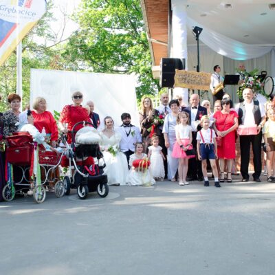 "Duża grupa dorosłych i dzieci pozująca na tle białej ścianki i sceny. Panna młoda w białej sukni z bukietem, pan młody w białym garniturze i czarnej muszce. Jeden z mężczyzn gra na akordeonie. W centralnej części grupy widać drewniany szyld z napisem: 'SOŁECTWO GRĘBOCICE'. Dzieci w jasnych strojach stoją po prawej stronie."