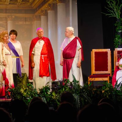 Aktorzy seniorzy na scenie teatralnej podczas spektaklu stylizowanego na starożytny Rzym. Pięć osób w białych togach i czerwonych lub fioletowych pelerynach stoi obok dekoracji z roślin, rozmawiając z kobietą w białej sukni i koronie, która siedzi na dużym krześle z czerwonym obiciem. W tle wyświetlany jest cyfrowy obraz antycznej sali z kolumnami.