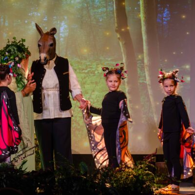 Scena ze spektaklu teatralnego. Dorosła osoba w masce osła, jasnej koszuli i kamizelce trzyma za ręce dwoje dzieci przebranych za motyle w czarnych strojach z kolorowymi skrzydłami. Po lewej stronie stoi postać w zielonym stroju i kwiecistym nakryciu głowy. W tle widać cyfrową projekcję lasu.