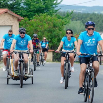 Czworo rowerzystów w błękitnych koszulkach z logo jedzie drogą w pobliżu domów. Dwoje mężczyzn na przedzie ma przypięte do rowerów przyczepki dziecięce. Tło to podmiejskie domy z czerwonymi dachami i pagórkowaty krajobraz.
