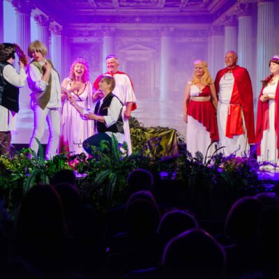 Grupa aktorów w różnym wieku na scenie podczas spektaklu teatralnego w rzymskich i historycznych strojach. Na pierwszym planie, pośrodku zielonych dekoracji, jedna z postaci klęczy przed kobietą w białej sukni. W tle widoczna jest fioletowo-różowa projekcja pałacowej sali z kolumnami, a z przodu zarysy głów publiczności siedzącej na widowni.