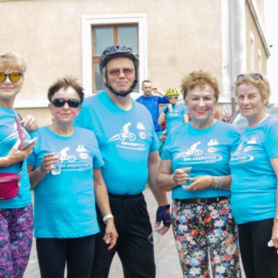 Pięć kobiet w identycznych błękitnych koszulkach z logo stoi razem, pozując do zdjęcia. Mają okulary przeciwsłoneczne. Dwie kobiety trzymają napoje w plastikowych kubkach. Tło to zabytkowy, jasny budynek, zaparkowane samochody i czarna, ozdobna latarnia uliczna.