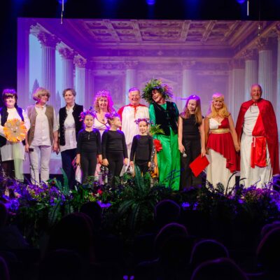 Wszyscy uczestnicy spektaklu teatralnego stoją w rzędzie na scenie, kłaniając się publiczności. Grupa składa się z dzieci w strojach motyli oraz dorosłych i seniorów w różnorodnych kostiumach – od antycznych tog po baśniowe szaty. Scena jest udekorowana roślinami, a w tle wyświetla się rzymska architektura.