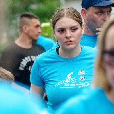 Bliski kadr na twarz kobiety z zaczesanymi do tyłu włosami, która patrzy w dół. Ma na sobie błękitną koszulkę z logo. Za nią, w rozmytym tle, widać inne osoby w podobnych koszulkach i zieloną roślinę.