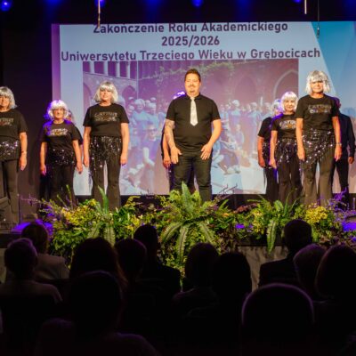 Uroczystość zakończenia roku akademickiego 2025/2026 Uniwersytetu Trzeciego Wieku w Grębocicach. Na scenie stoi mężczyzna w czarnej koszulce i krawacie, otoczony przez grupę kobiet w błyszczących, srebrnych perukach, czarnych koszulkach i ciemnych spodniach z cekinami. W tle widoczny jest ekran z napisem informacyjnym oraz zdjęciem uczestników.