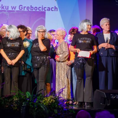 Grupa seniorek w srebrnych perukach i czarnych koszulkach z napisem „Latino Star" stoi na scenie obok elegancko ubranych organizatorów. Jedna z kobiet trzyma w rękach dyplom. Po prawej stronie mężczyzna w garniturze mówi do mikrofonu, a obok niego stoi kobieta w granatowej garsonce.