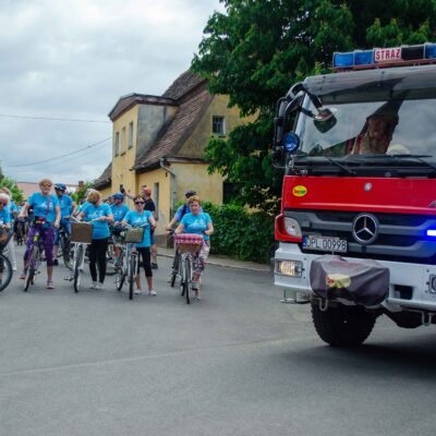 Duży, czerwony wóz strażacki Mercedes-Benz z napisem "OSP KWELICE" jedzie na czele kolumny, z włączonymi kogutami. Za nim jedzie mniejsza grupa rowerzystów w błękitnych koszulkach z logo. Tło to wiejska droga, zieleń i zabytkowy budynek.